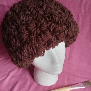 May include: A brown crocheted hat with a textured, looped design. The hat is displayed on a white mannequin head against a pink background. A paintbrush with a wooden handle and a blue tip rests below the head.