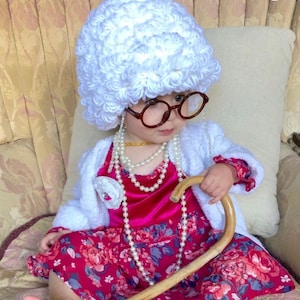 Puede incluir: Un niño pequeño vestido como una mujer mayor que lleva una peluca blanca rizada, gafas redondas, un vestido rosa con estampado floral, una chaqueta blanca, un collar de perlas y un bastón de madera.