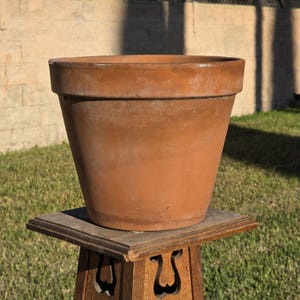 May include: A terracotta plant pot with a wide, thick rim and a tapered body. The surface of the pot is a matte, earthy orange-brown color with subtle variations in tone and texture. The rim features a faint, embossed stamp reading Made in Italy.
