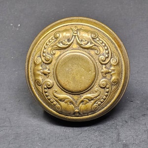 May include: A round, brass door knob with an ornate, floral design. The knob has a raised, circular centre.