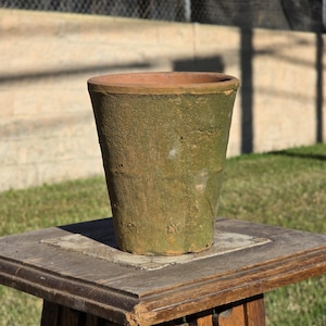 May include: A tapered terracotta plant pot with a weathered, mossy green exterior finish and a smooth, unglazed orange-brown rim. The surface of the vessel shows a textured, aged appearance with visible imperfections and earthy color variations.