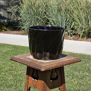 May include: A glossy black ceramic planter with a spiral design sits on a wooden plant stand. The planter is cylindrical with a wide opening. The stand is made of wood and has decorative cutouts. The background features green grass and foliage.
