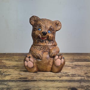 May include: A brown ceramic bear cookie jar featuring a textured fur finish, blue eyes, a black nose, and a small red tongue visible. The bear is in a seated position with its front paws resting on its belly and light pink paw pads on its feet.