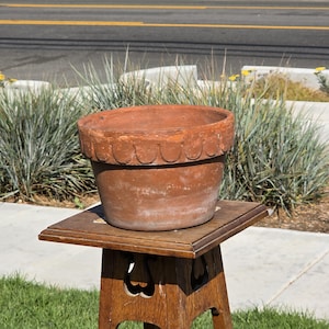 May include: A weathered, terracotta-colored flower pot with a scalloped rim sits on a wooden plant stand. The pot has a wide opening and a slightly textured surface. The stand is made of wood and has a decorative cutout design.