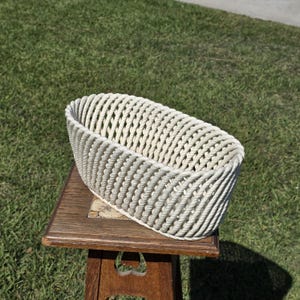 Puede incluir: Una cesta ovalada tejida de color blanquecino. La cesta está hecha de cuerda gruesa y retorcida, creando una superficie texturizada. La cesta está sobre una mesa de madera con una tapa de azulejos decorativos, sobre un fondo de hierba verde.