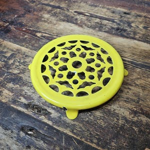 May include: Yellow metal trivet with a decorative, circular, openwork design. The trivet has three short legs.