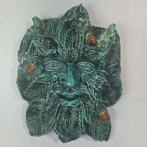 May include: A green ceramic wall plaque depicting a face formed from layered leaves. The sculpture features a central face with detailed eyes, a nose, and a leafy beard, accented by three small brown acorns nestled among the foliage.