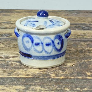 May include: A small, ceramic jar with a lid, featuring a blue and white floral design. The jar has a round shape with two small handles and a blue knob on the lid. The jar's exterior is decorated with blue abstract patterns.