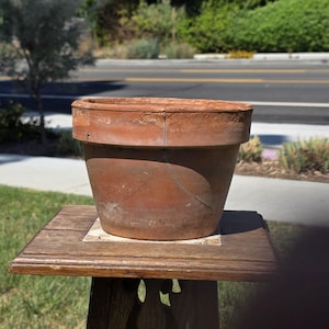 May include: A weathered terracotta flower pot with a wide, thick rim and a visible vertical crack extending from the top edge down the side. The clay surface displays a rustic, matte texture with natural color variations in shades of brown.