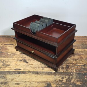 May include: A dark brown wooden jewelry organizer featuring a top tray with four grey velvet watch cushions, an adjacent open compartment, a middle open shelf, and a bottom drawer with a small round brass knob for storage and organization.
