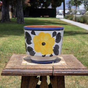 May include: A ceramic planter featuring a white base with a hand-painted yellow flower and black leaf motifs. The pot has a wide, flared rim and a tapered base, accented by horizontal blue bands at the top and bottom edges, with a terracotta interior.