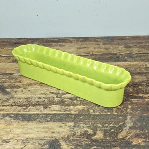 May include: A bright lime green rectangular ceramic planter with a scalloped rim. The planter is empty and sits on a weathered wooden surface. The planter is designed for flowers or herbs.