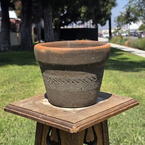 May include: A dark brown, weathered clay pot with a wide, circular opening and a thick, rounded rim. The exterior surface features a textured, horizontal band of incised, repeating diagonal line patterns encircling the middle of the vessel.