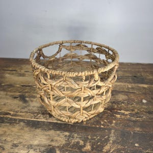 May include: A round, woven basket with a natural, light brown color. The basket has a decorative, open weave pattern around the top and a sturdy, woven body. The basket sits on a wooden surface.