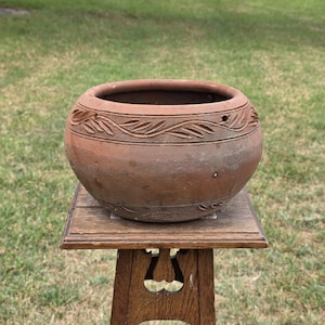 May include: A round, terracotta-colored clay bowl featuring a wide, open top and a textured, carved leaf pattern encircling the upper rim. The bowl has a matte, earthy finish with visible surface variations and sits on a wooden stand.