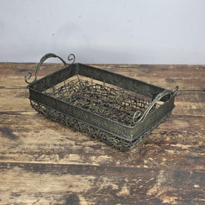 May include: A rectangular metal basket with a wire mesh interior and decorative grape vine pattern around the rim. The basket has curved handles and a distressed, antique finish, perfect for home decor.