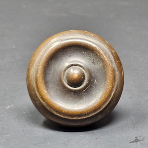 Puede incluir: Un pomo de puerta redondo de latón con un centro elevado y círculos concéntricos.