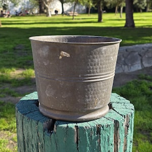 May include: A weathered, antique metal bucket with a hammered texture. The bucket is a dark, muted bronze color and sits on a green-painted wooden post. The background shows a grassy area and trees.