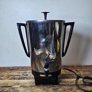 May include: A silver electric coffee percolator with a black handle and spout. The percolator has a black base and a cord.