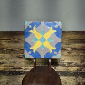 Puede incluir: Azulejo cerámico cuadrado con diseño floral. El azulejo presenta un fondo gris con motivos florales azules, amarillos y turquesas. El azulejo se exhibe sobre un soporte de madera oscura.