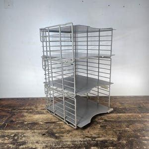 May include: A vintage metal desk organizer with four tiers. The organizer is made of gray metal with a wireframe design and solid shelves. The unit is designed to hold papers and documents. The organizer is sitting on a wooden surface.