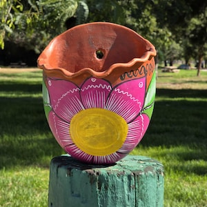 May include: Hand-painted ceramic wall planter with a vibrant floral design. The planter features a pink flower with a yellow center, green leaves, and a terracotta-colored rim. The decorative piece is mounted on a weathered wooden post.