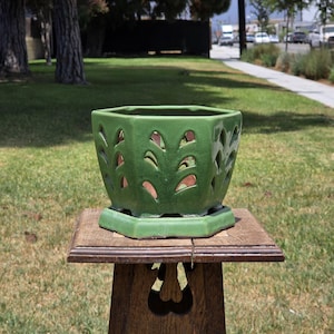 May include: A hexagonal green ceramic planter featuring a series of cut-out leaf-shaped patterns along its sides. The planter sits on a matching green ceramic base and has a smooth, glossy finish that reflects light across its structured surface.