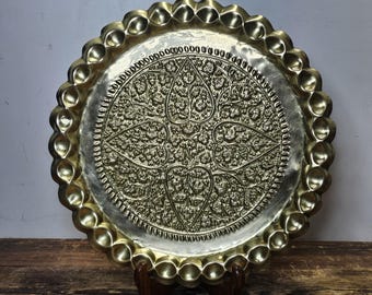 May include: A round, gold-colored metal tray with a decorative, embossed design of leaves and flowers. The tray has a scalloped edge.
