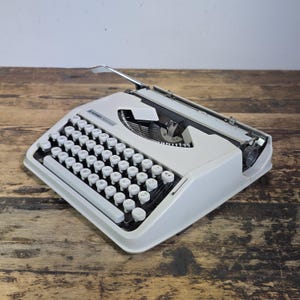 Może przedstawiać: Jasnoszara, przenośna maszyna do pisania Hermes Baby z klawiaturą QWERTY, białymi okrągłymi klawiszami, metalową dźwignią powrotu karetki i czarnym mechanizmem taśmy. Nazwa marki Hermes Baby jest nadrukowana na czarno na obudowie.
