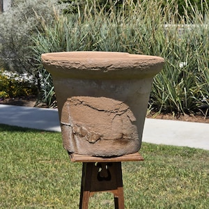 May include: A large, weathered terracotta planter with a wide, thick rim and a tapered body. The surface features a rough, textured finish with visible cracks and patches of discoloration, resting on a dark wooden stand with cutout details.