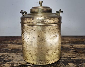 Puede incluir: Un recipiente de latón vintage con tapa y dos asas. El recipiente tiene un acabado martillado y está ligeramente deslustrado.