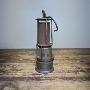 May include: An antique metal miner's lamp with a glass enclosure and a handle. The lamp has a cylindrical shape with a rusty brown fabric covering the light source. The base and top are made of metal, and the lamp sits on a wooden surface.