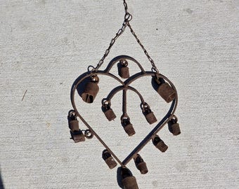Könnte beinhalten: Ein herzförmiger Wandbehang aus Metall mit rostiger Oberfläche und einer Reihe kleiner, hängender Glocken, die mit dünnen Metallketten am Rahmen befestigt sind. Die Struktur enthält oben eine zentrale Kettenschlaufe zum Aufhängen.