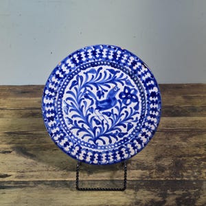 May include: A round ceramic plate featuring a white base with hand-painted cobalt blue designs. The center displays a bird surrounded by leafy foliage and a flower, while the rim is decorated with a repeating geometric pattern of blue arches.