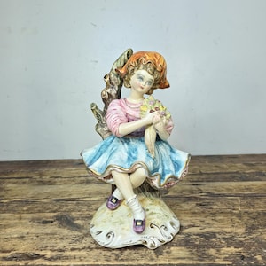 May include: A porcelain figurine of a seated girl holding flowers. She wears a pink top, a blue skirt, and a red hat. The figurine is perched on a decorative base with gold accents. The girl's shoes are purple.