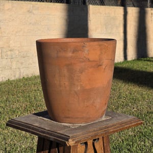 May include: A terracotta plant pot with a tapered cylindrical shape and a smooth, matte finish. The pot features natural color variations in shades of reddish-brown, showing a weathered texture across the exterior surface.