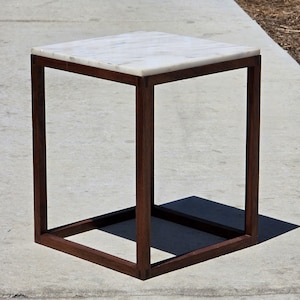 May include: A square side table featuring a white marble top with subtle grey veining. The base is constructed from four dark brown wooden legs connected by horizontal wooden supports at the bottom, forming a clean, open, and geometric frame.