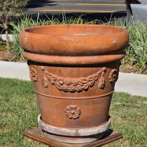 May include: A terracotta plant pot featuring a wide, rounded rim and a tapered body. The exterior is decorated with relief carvings of floral swags and individual flower motifs. The pot sits on a matching circular base with a light-colored edge.