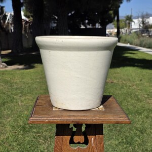 Peut inclure: Un pot de fleur en céramique crème avec un bord légèrement évasé. Le pot repose sur un support de plante en bois avec un dessus carré et des découpes décoratives. Le support est en bois foncé.