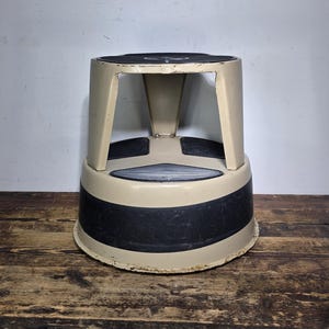 May include: A beige and black step stool with a circular top and base. The step stool has a black band around the base and black non-slip pads on the top. The step stool is made of metal and is on a wooden surface.