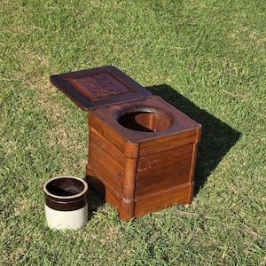 May include: A vintage wooden commode with a hinged lid, featuring carved floral details, and a round opening. A small, cylindrical stoneware chamber pot with a brown band sits beside it on the grass.