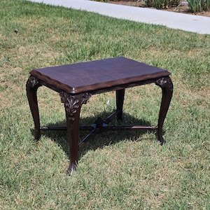 May include: A dark brown wooden side table with a rectangular top featuring wavy edges. The table stands on four curved legs, each adorned with carved leaf-like details at the top, and includes a central cross-shaped stretcher connecting the legs.