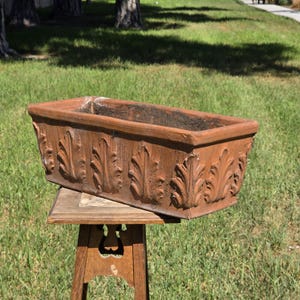 May include: A rectangular terracotta planter featuring a repeating embossed leaf pattern along the exterior sides. The planter has a wide, open top and a weathered, earthy orange-brown finish, resting on a wooden stand.