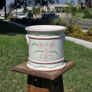 May include: A white ceramic planter with a cylindrical shape, featuring hand-painted pink roses, green foliage, and a pink bow motif. The rim and base are decorated with a raised pink rope-like texture, and small handles are on the sides.