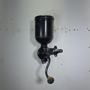 Op de afbeelding: Een aan de muur gemonteerde zwarte metalen koffiemolen met een cilindrische trechter, een slinger met een houten knop en een montagebeugel met de in reliëf aangebrachte tekst "REGINA". Het apparaat heeft een matzwarte afwerking en een gebogen hendel.
