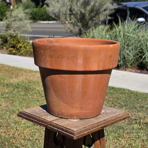 May include: A terracotta plant pot with a wide, flat rim and a tapered body. The surface of the clay is smooth with natural color variations in shades of reddish-brown, featuring a distinct horizontal band around the upper section.