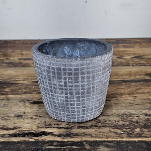 May include: A gray, textured, small plant pot with a grid pattern. The pot is made of a concrete-like material and has a round shape. The pot is sitting on a wooden surface.