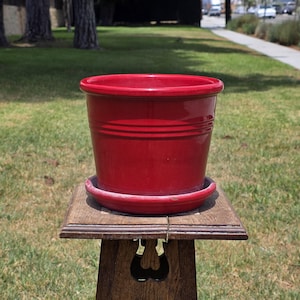 May include: A glossy, solid red ceramic plant pot with a matching circular saucer base. The pot features a simple, cylindrical shape with a slightly flared rim and a subtle horizontal grooved line detail encircling the upper exterior surface.