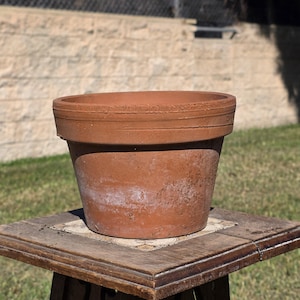May include: A terracotta plant pot with a wide, thick rim and a tapered body. The surface of the clay is reddish-brown with visible natural texture, slight weathering, and subtle variations in colour throughout the exterior of the vessel.