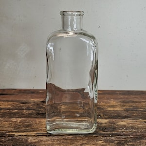 Op de afbeelding: Een heldere glazen fles met een vierkante vorm en een afgeronde bovenkant. De fles is leeg en heeft een glad oppervlak.